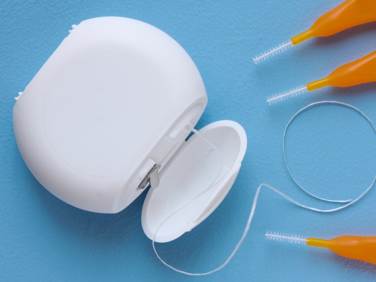 Filo interdentale e scovolini di misure diverse per la pulizia degli spazi fra i denti, appoggiati su fondo azzurro.