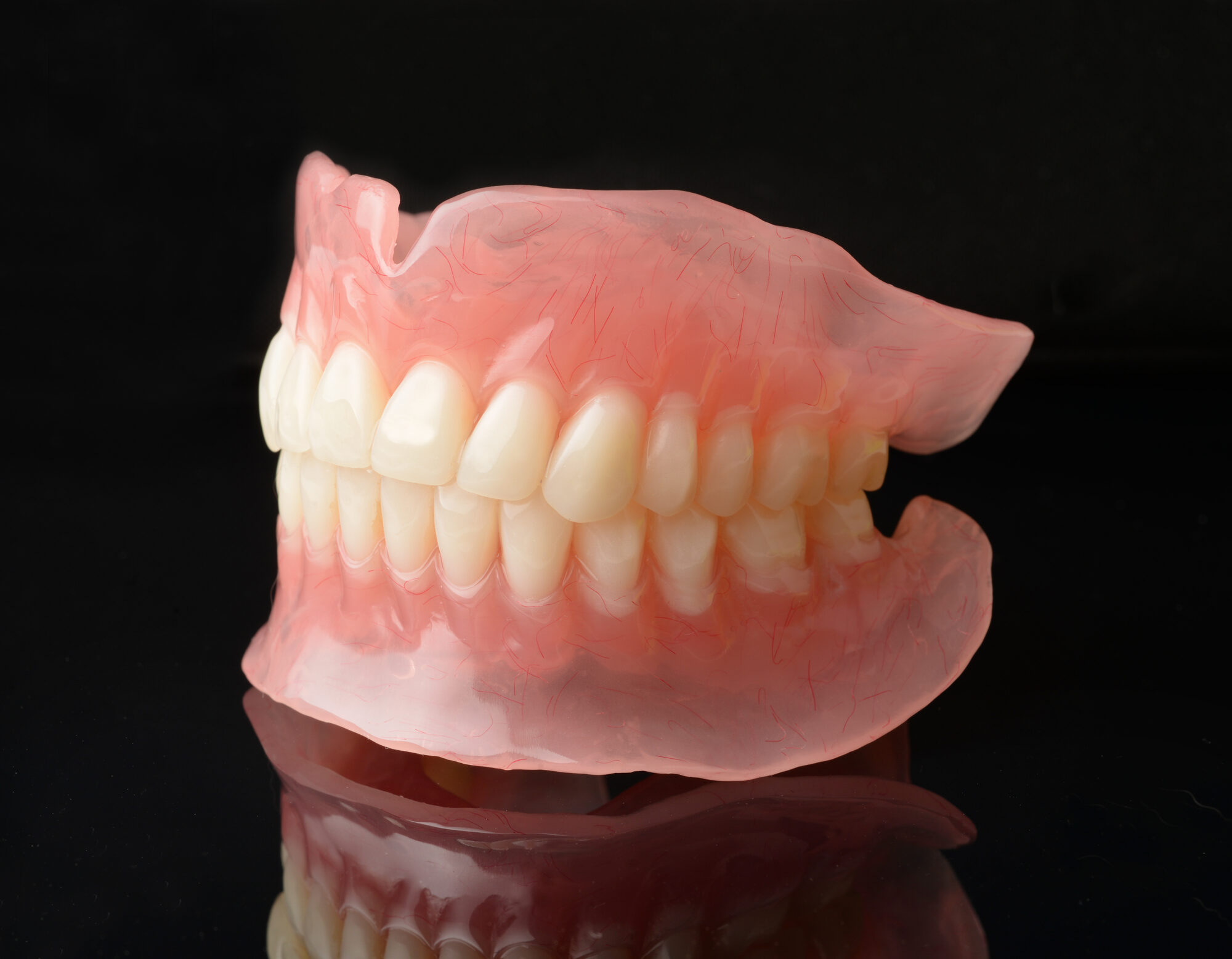 Protesi dentale totale superiore e inferiore in resina rosa con denti avorio, fotografata su fondo nero riflettente