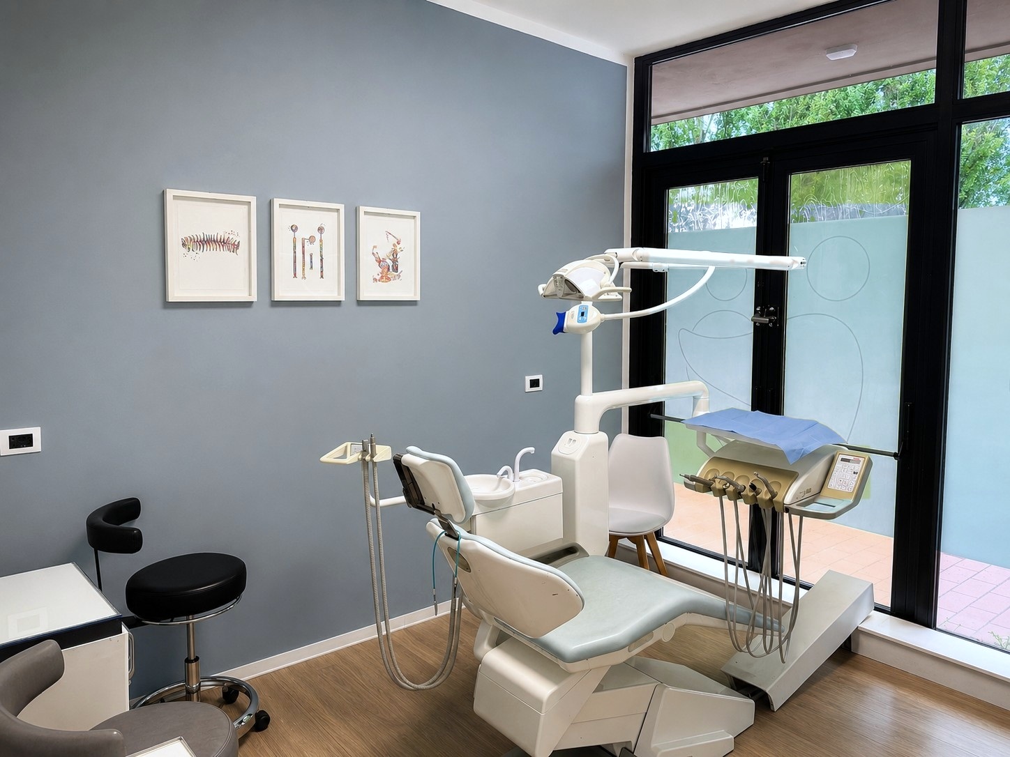 Sala operativa dello Studio Dentistico Lera con parete grigia, stampe incorniciate e ampia vetrata sul verde
